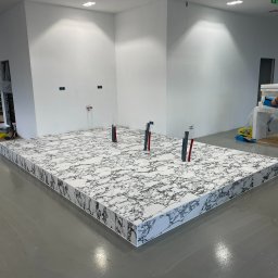 CREO JAKUB CAŁA - Podwyższona podłoga wykończona płytami imitującymi marmur w surowym wnętrzu. Widoczne wystające rury instalacyjne i gładka, szara posadzka.