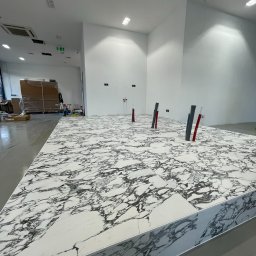 CREO JAKUB CAŁA - Biała marmurowa posadzka z widocznymi pionami instalacji w minimalistycznym wnętrzu. Widok z perspektywy podłogi, jasne oświetlenie.