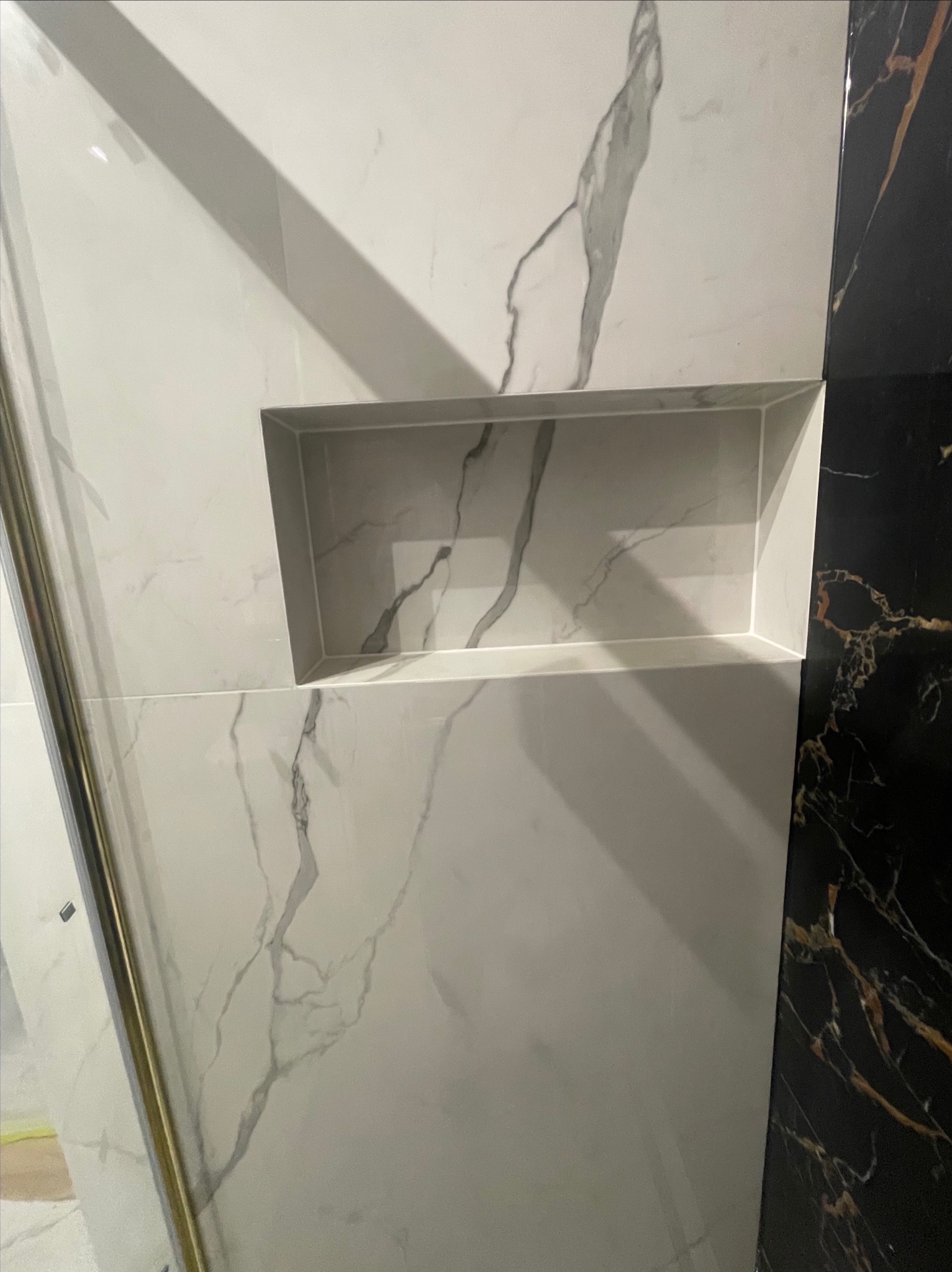 Ściana wyłożona białymi płytkami imitującymi marmur z wnęką, obok fragment ściany z czarnymi płytkami z marmurkowym wzorem.