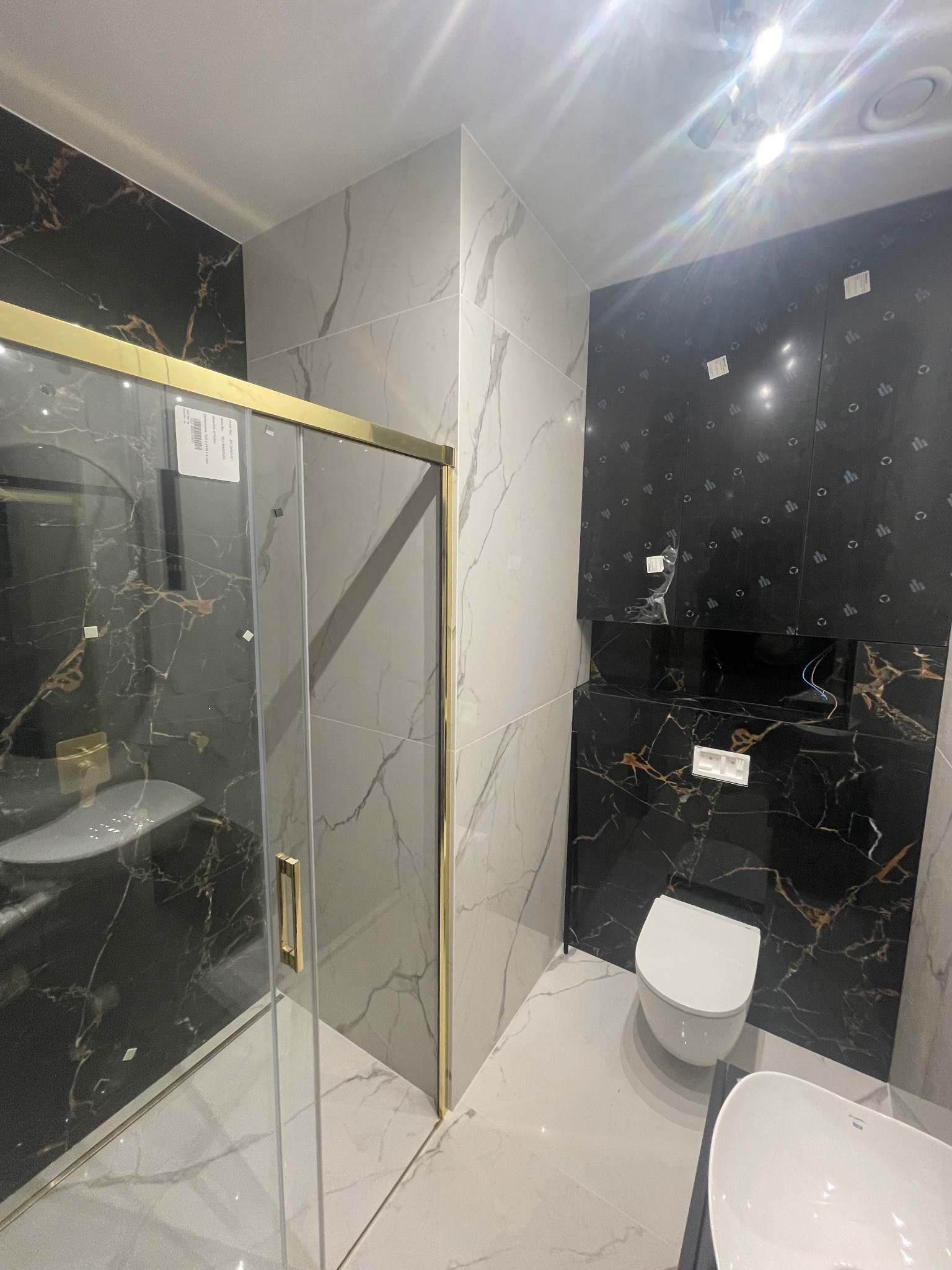 Nowoczesna łazienka z kabiną prysznicową o złotych profilach, marmurowymi płytkami w odcieniach czerni i bieli, wiszącą toaletą i umywalką nablatową.