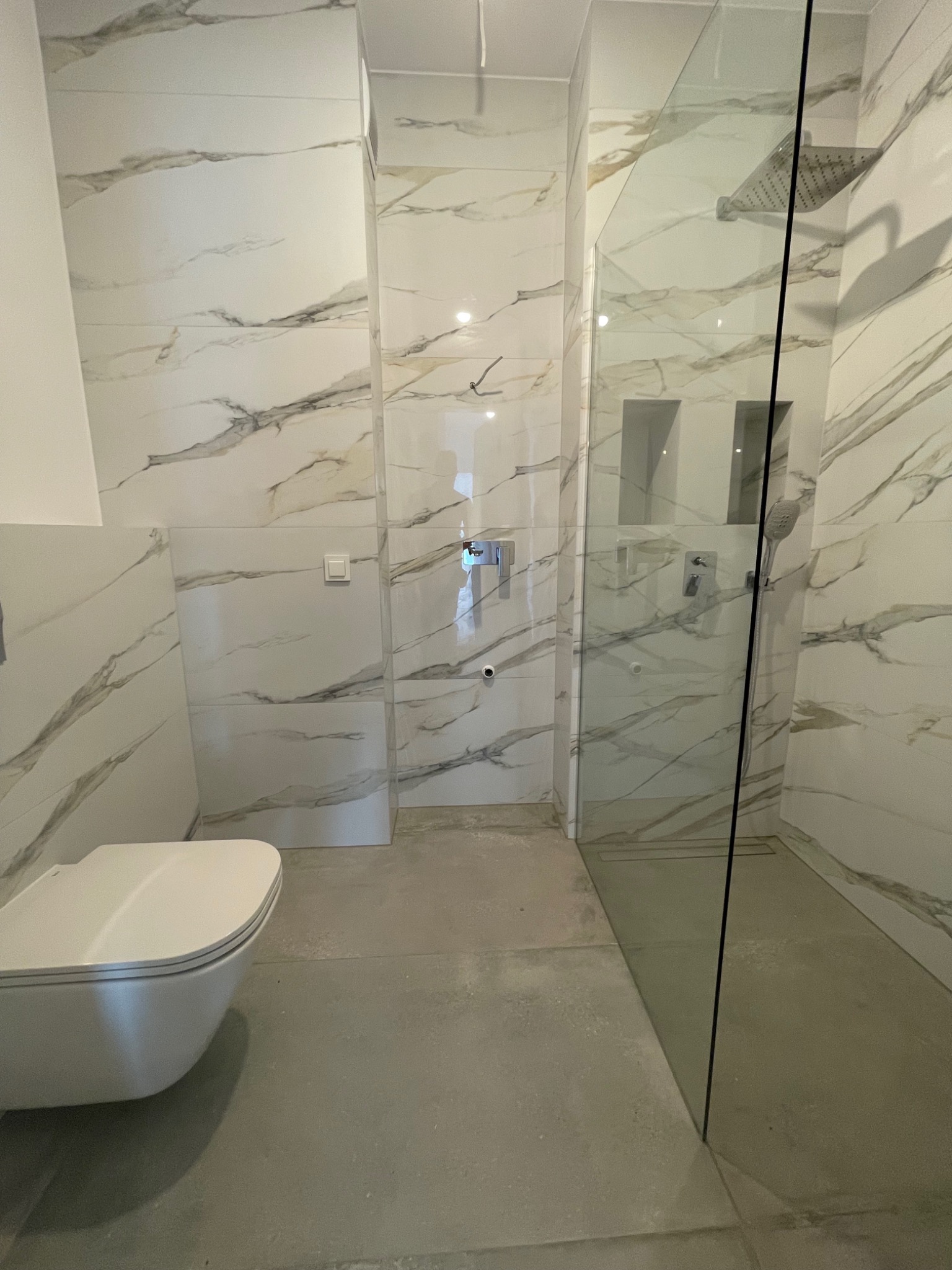 Nowoczesna łazienka z białymi płytkami imitującymi marmur, kabiną prysznicową typu walk-in z czarnymi elementami, podtynkową baterią, wiszącą toaletą i szarą podłogą.