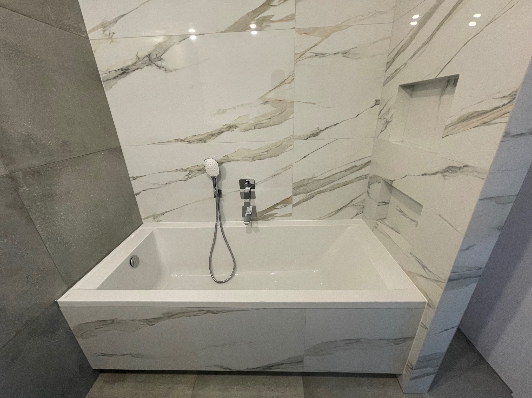 Nowoczesna łazienka z białą wanną obudowaną marmurkowymi płytkami, szara ściana z płytek po lewej stronie, wnęka w ścianie nad wanną.