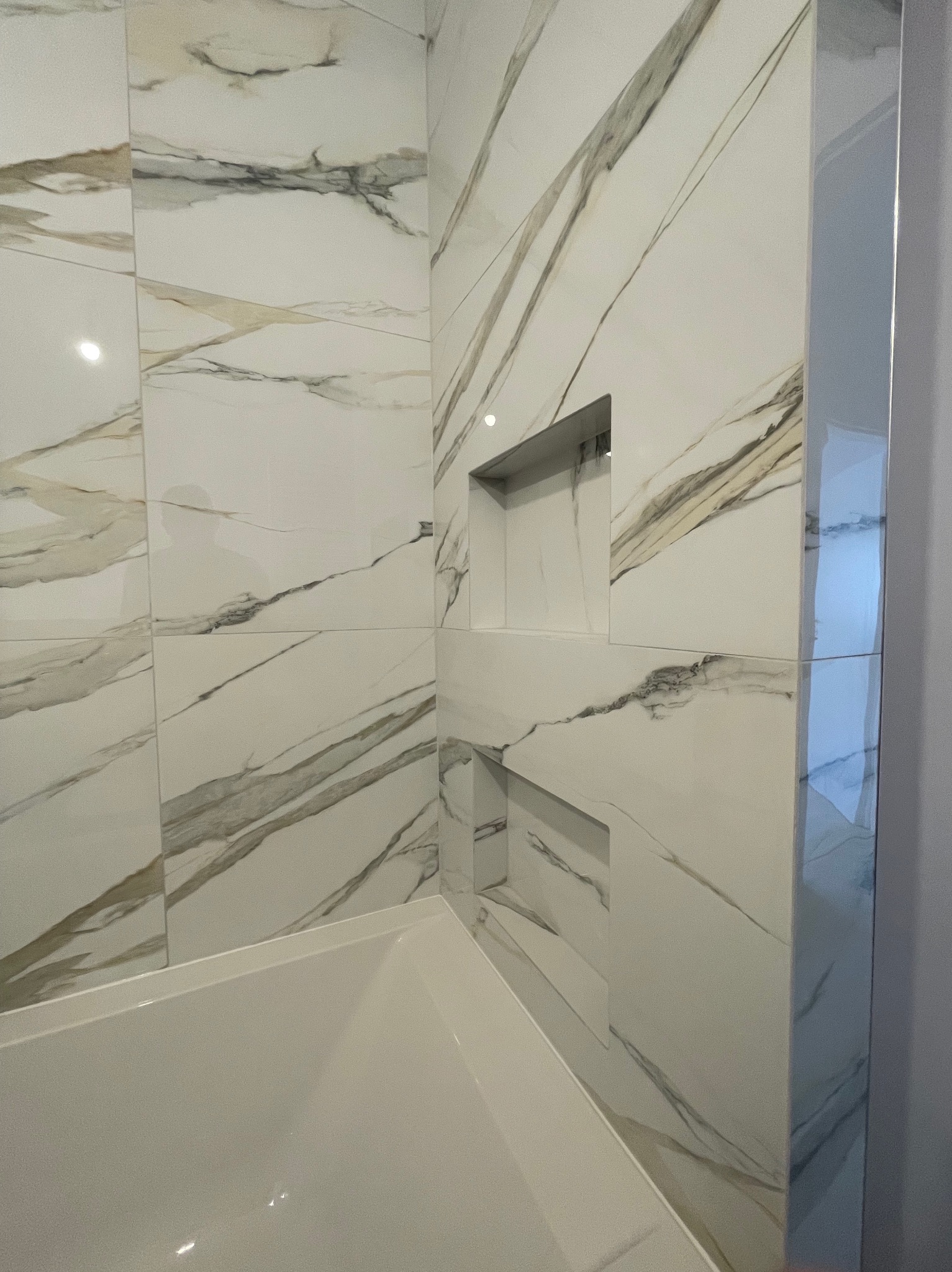 Białe płytki imitujące marmur z szarymi i złotymi żyłkami, wbudowane półki w strefie wanny, wykończenie narożników listwą metalową.