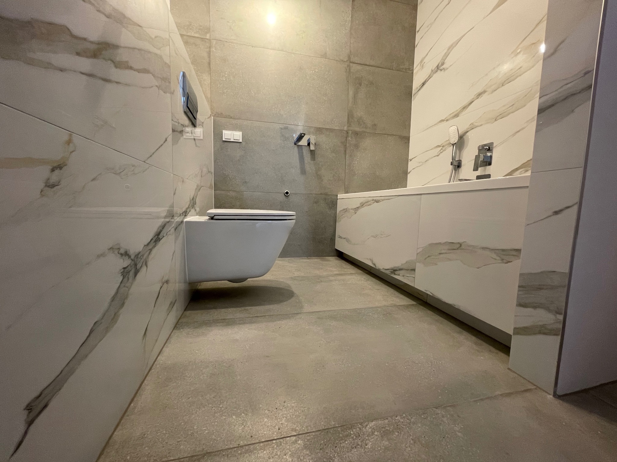 Nowoczesna łazienka z podwieszaną toaletą, szarymi płytkami imitującymi beton na ścianie i marmurowymi płytkami z białym tłem i szarymi żyłami na pozostałych ścianach i zabudowie wanny.