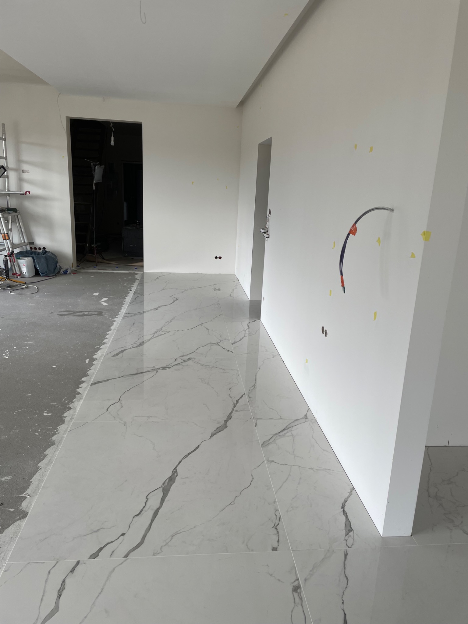 Układanie płytek imitujących marmur w nowym mieszkaniu, widoczne przygotowane ściany i surowa posadzka w sąsiednim pomieszczeniu, drabina i narzędzia w tle.