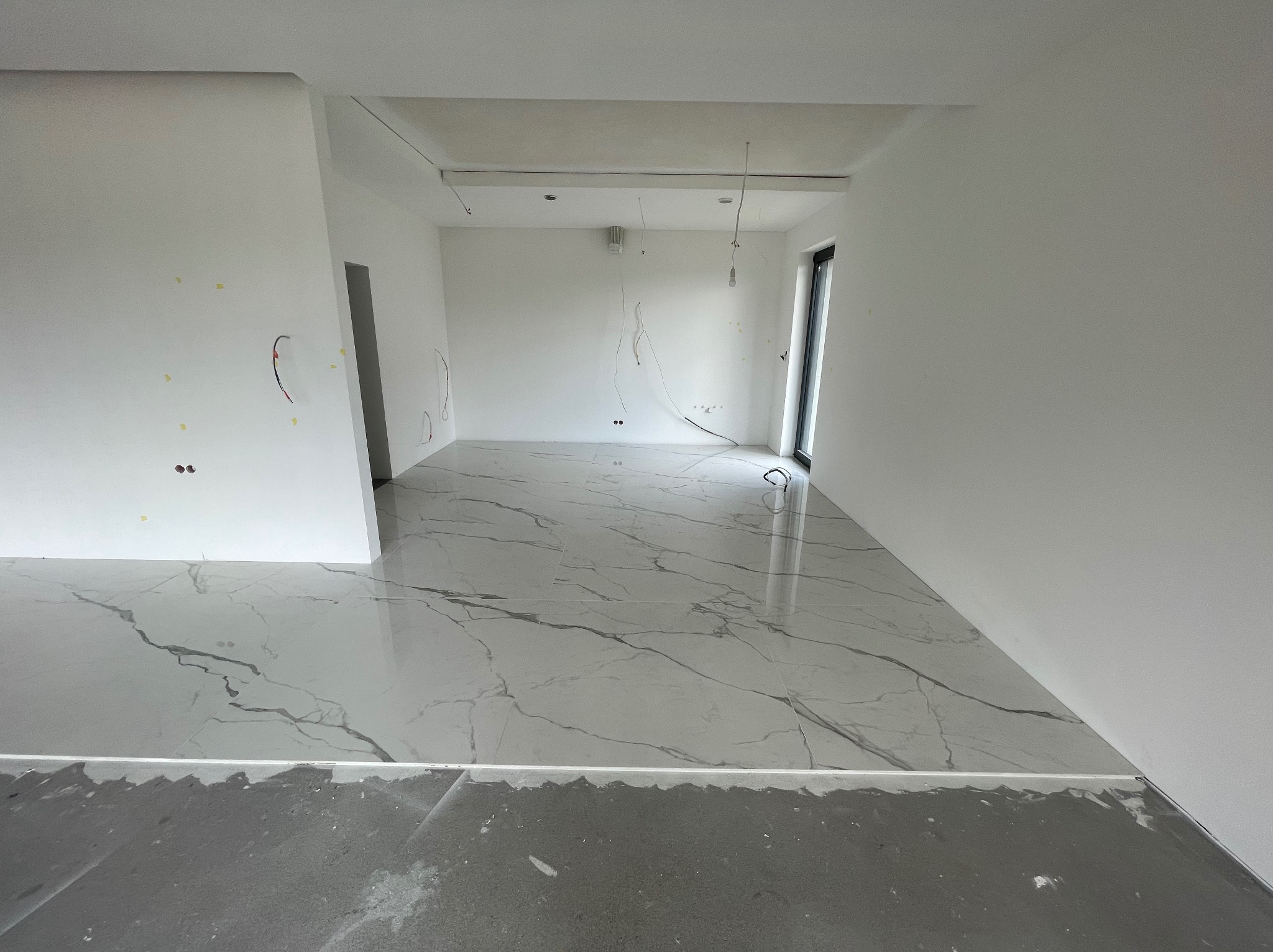 Nowoczesne wnętrze w trakcie prac wykończeniowych: białe ściany z widocznymi przewodami elektrycznymi, marmurowa podłoga z szarymi żyłami i surowa wylewka przy ścianach.