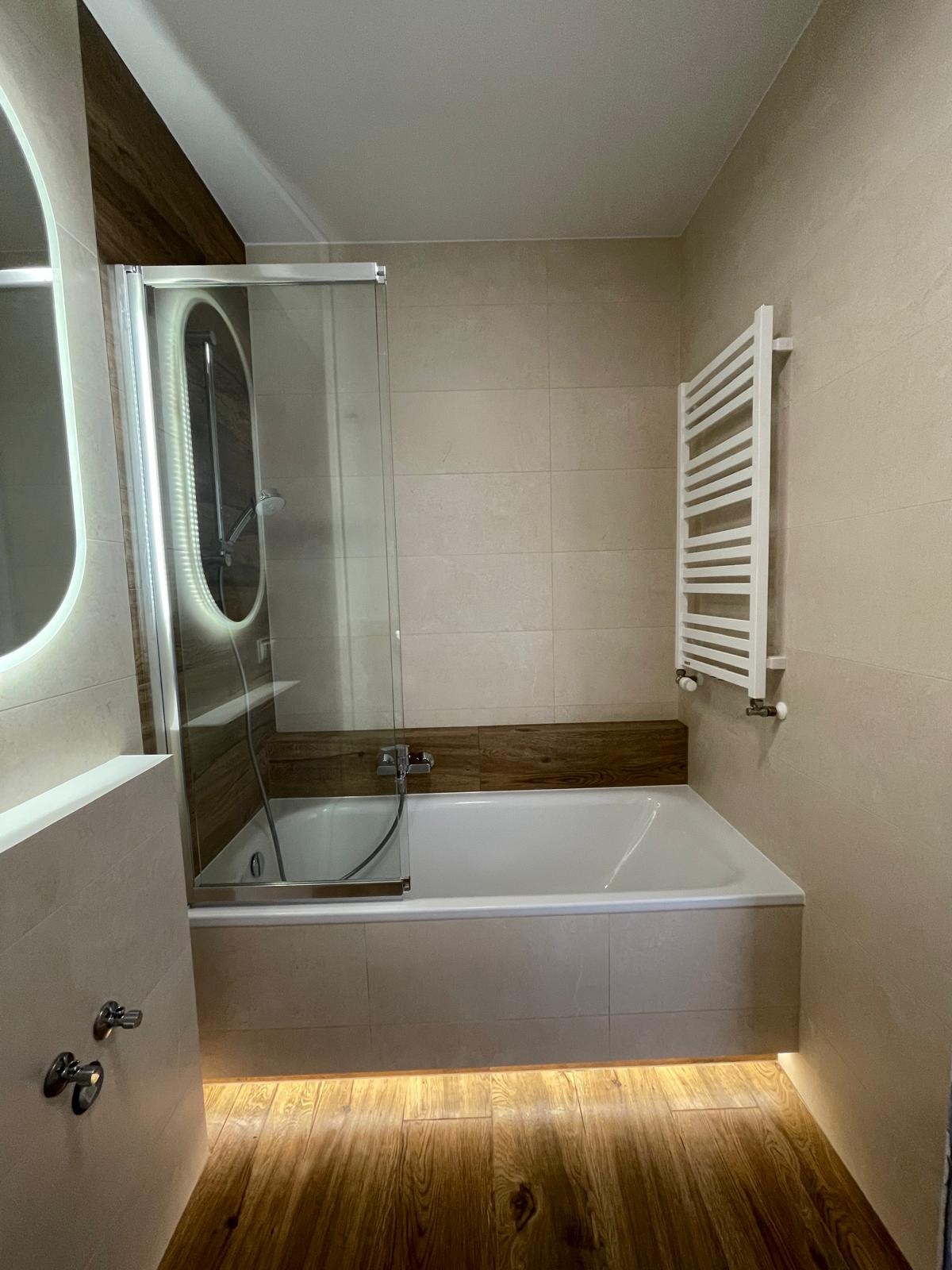 Jasna łazienka z wanną obudowaną płytkami, szklanym parawanem, grzejnikiem drabinkowym i podświetlaną podłogą z paneli drewnopodobnych.