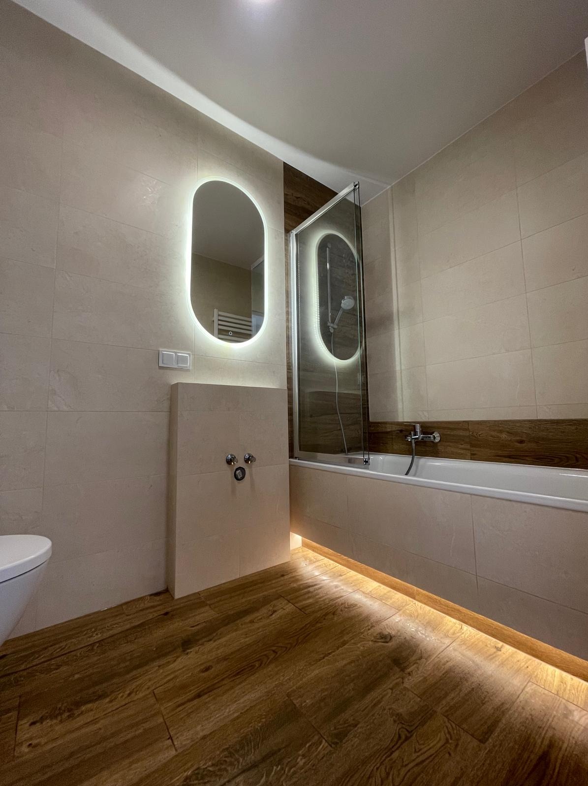 Nowoczesna łazienka z podświetlanymi lustrami i drewnopodobną podłogą. Kabina prysznicowa z szybą i chromowaną armaturą. Beżowe płytki na ścianach.
