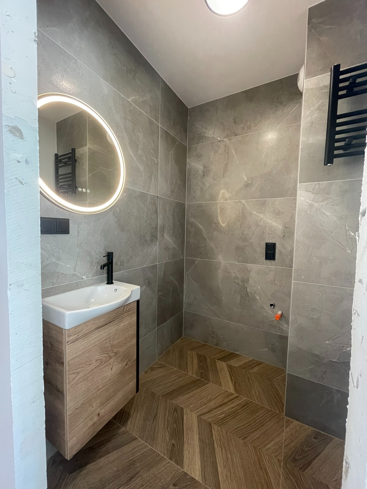 Nowoczesna łazienka z szarymi płytkami imitującymi marmur, drewnianą szafką pod umywalką i podłogą ułożoną w jodełkę. Okrągłe lustro z podświetleniem LED.