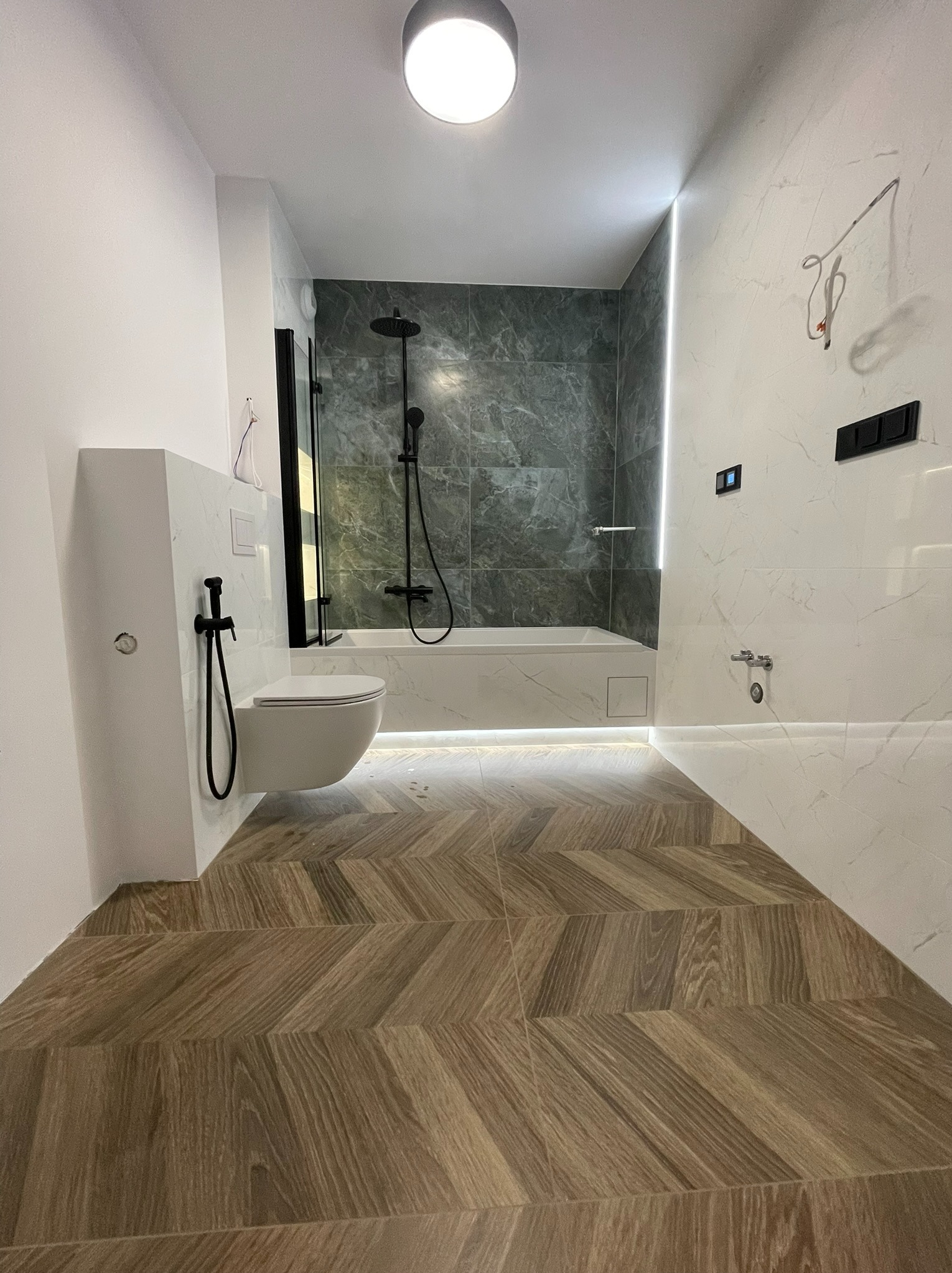 Nowoczesna łazienka po remoncie z drewnopodobną podłogą ułożoną w jodełkę, białe marmurowe płytki na ścianach i czarna armatura.