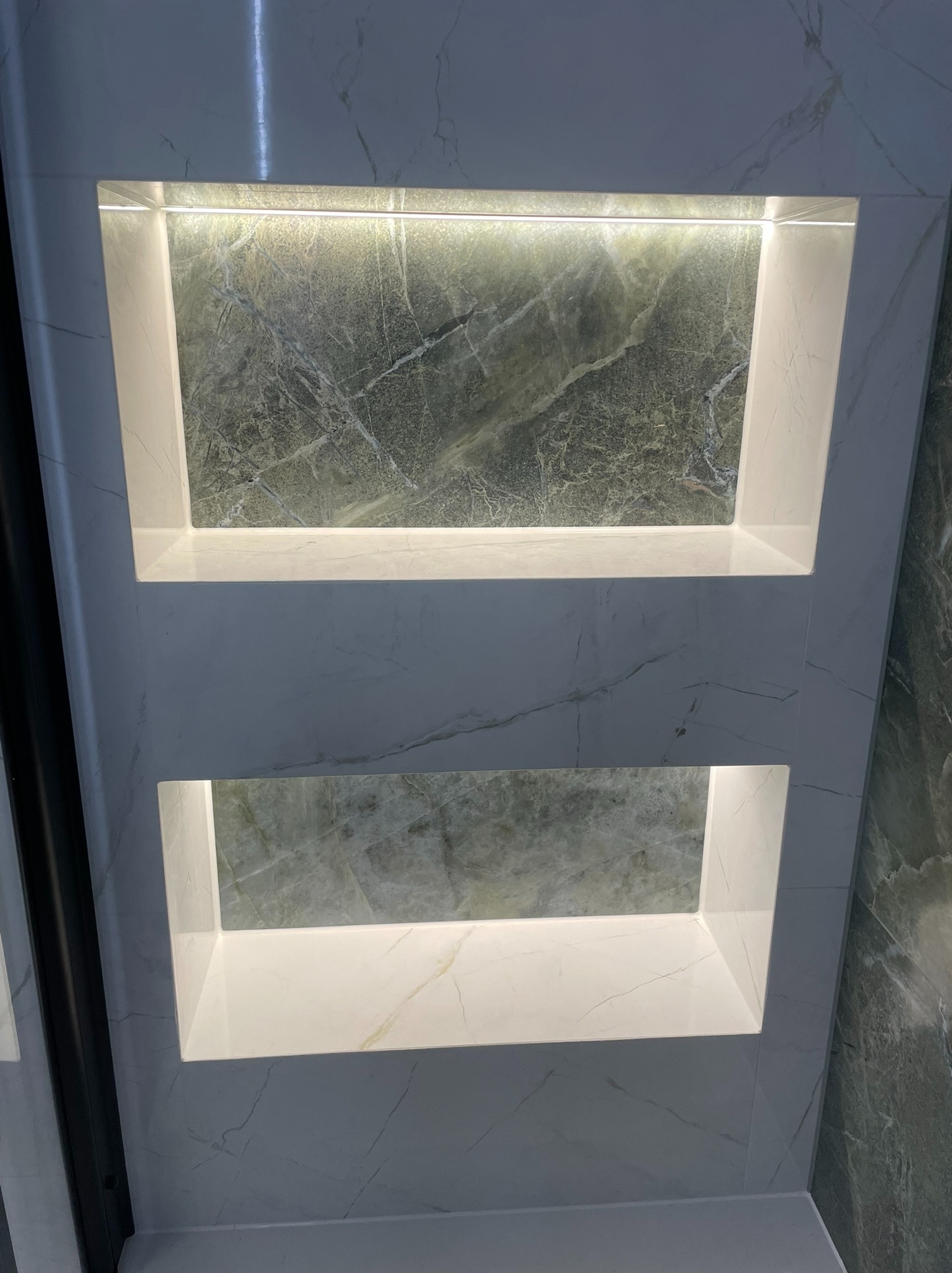 Dwa podświetlane wnęki w ścianie wyłożonej płytkami imitującymi marmur. Górna wnęka z zielonym marmurem, dolna z białym. Nowoczesny detal wykończenia łazienki.