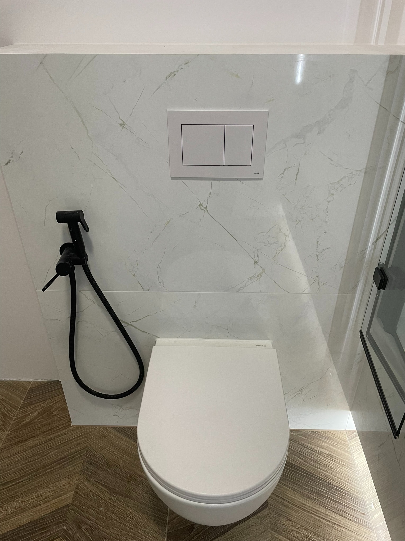 Nowoczesna toaleta wisząca z czarnym bidetem i marmurowymi płytkami. Widoczna deska sedesowa i przycisk spłukiwania. Podłoga wyłożona panelami imitującymi drewno.