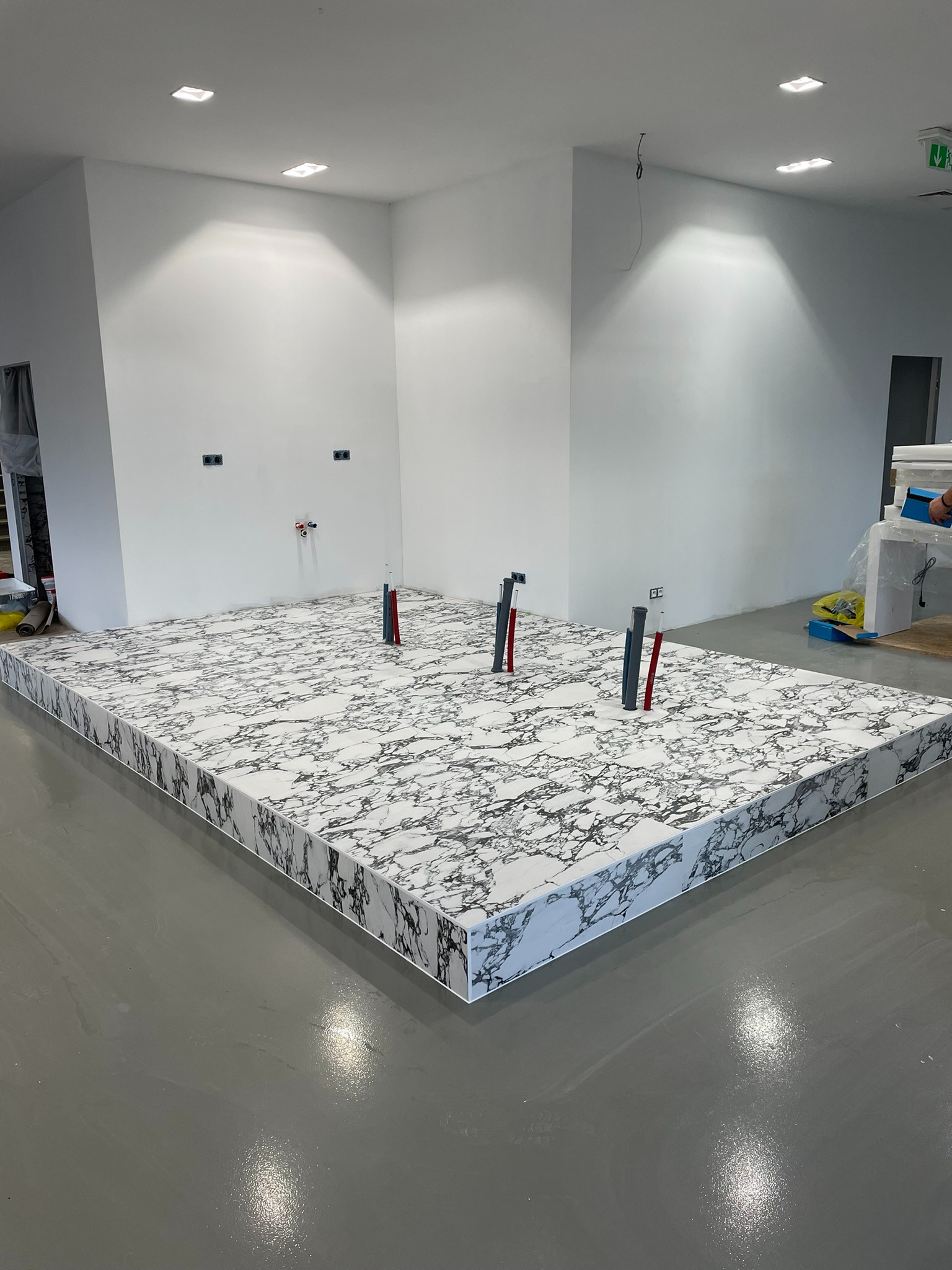 Podwyższona podłoga wykończona płytami imitującymi marmur w surowym wnętrzu. Widoczne wystające rury instalacyjne i gładka, szara posadzka.