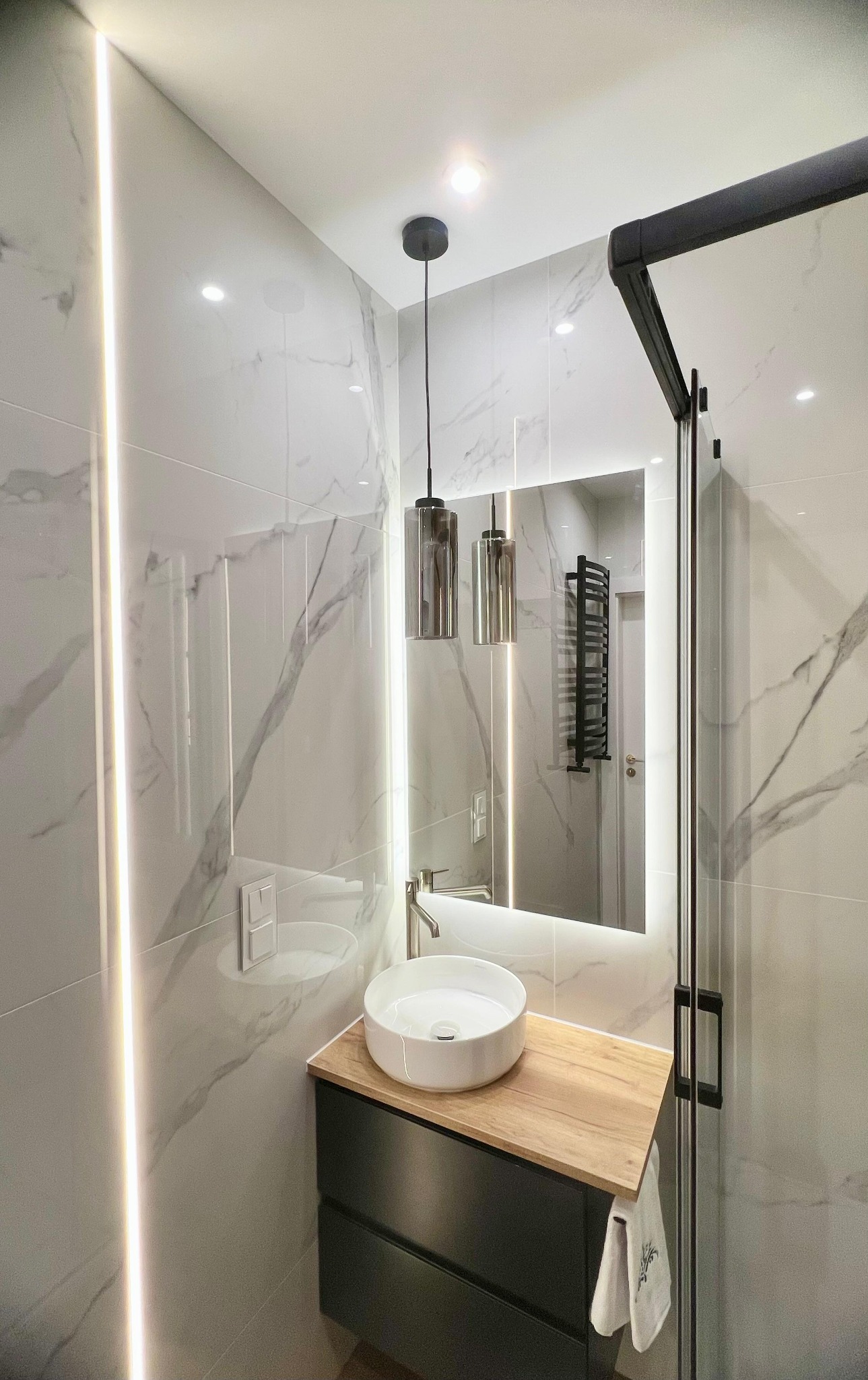 Nowoczesna łazienka z marmurowymi płytkami, podświetlanym lustrem, okrągłą umywalką na drewnianym blacie i czarną kabiną prysznicową.