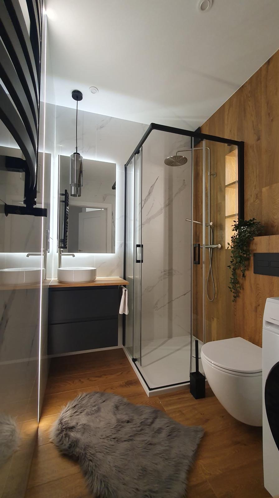 Nowoczesna łazienka z kabiną prysznicową typu walk-in, umywalką na szafce i drewnianą okładziną ściany. Widoczny grzejnik drabinkowy i puszysty dywanik.