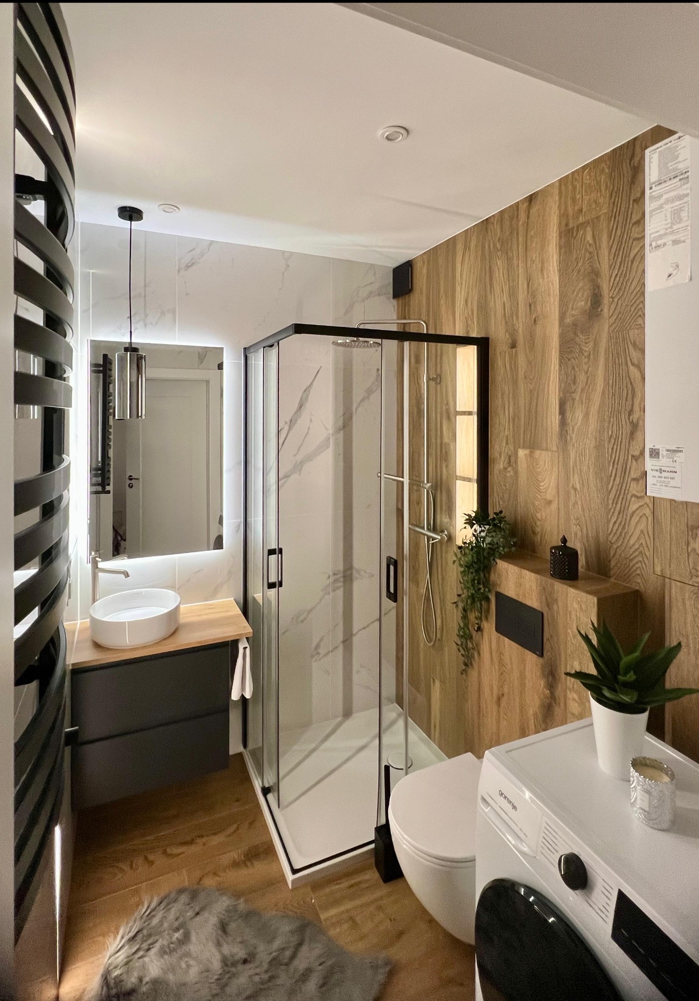 Nowoczesna łazienka z drewnianymi i marmurowymi akcentami, kabiną prysznicową typu walk-in i okrągłą umywalką nablatową. Uwagę zwraca grzejnik dekoracyjny.