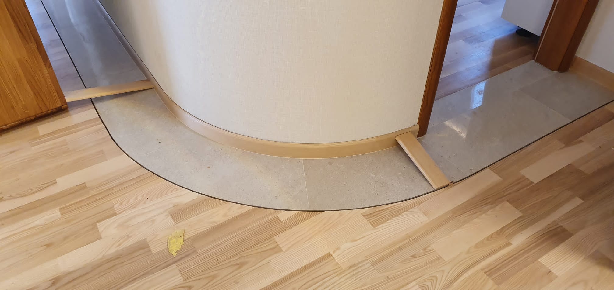 Fragment podłogi z ułożonymi panelami w jasnym odcieniu drewna, przechodzącymi w szarą, kamienną posadzkę wzdłuż zakrzywionej ściany, wykończonej drewnianą listwą przypodłogową, z widocznym...