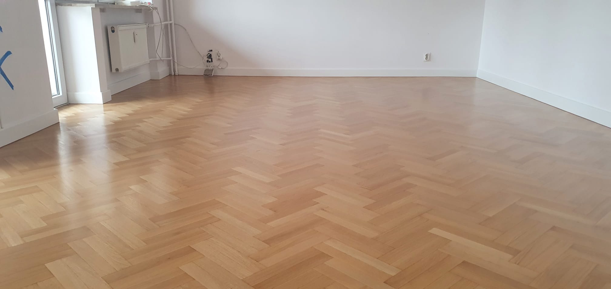 Jasny, odnowiony parkiet ułożony we wzór jodełki francuskiej, widok na puste pomieszczenie z białymi ścianami i listwami przypodłogowymi, grzejnik i gniazdko elektryczne przy ścianie.