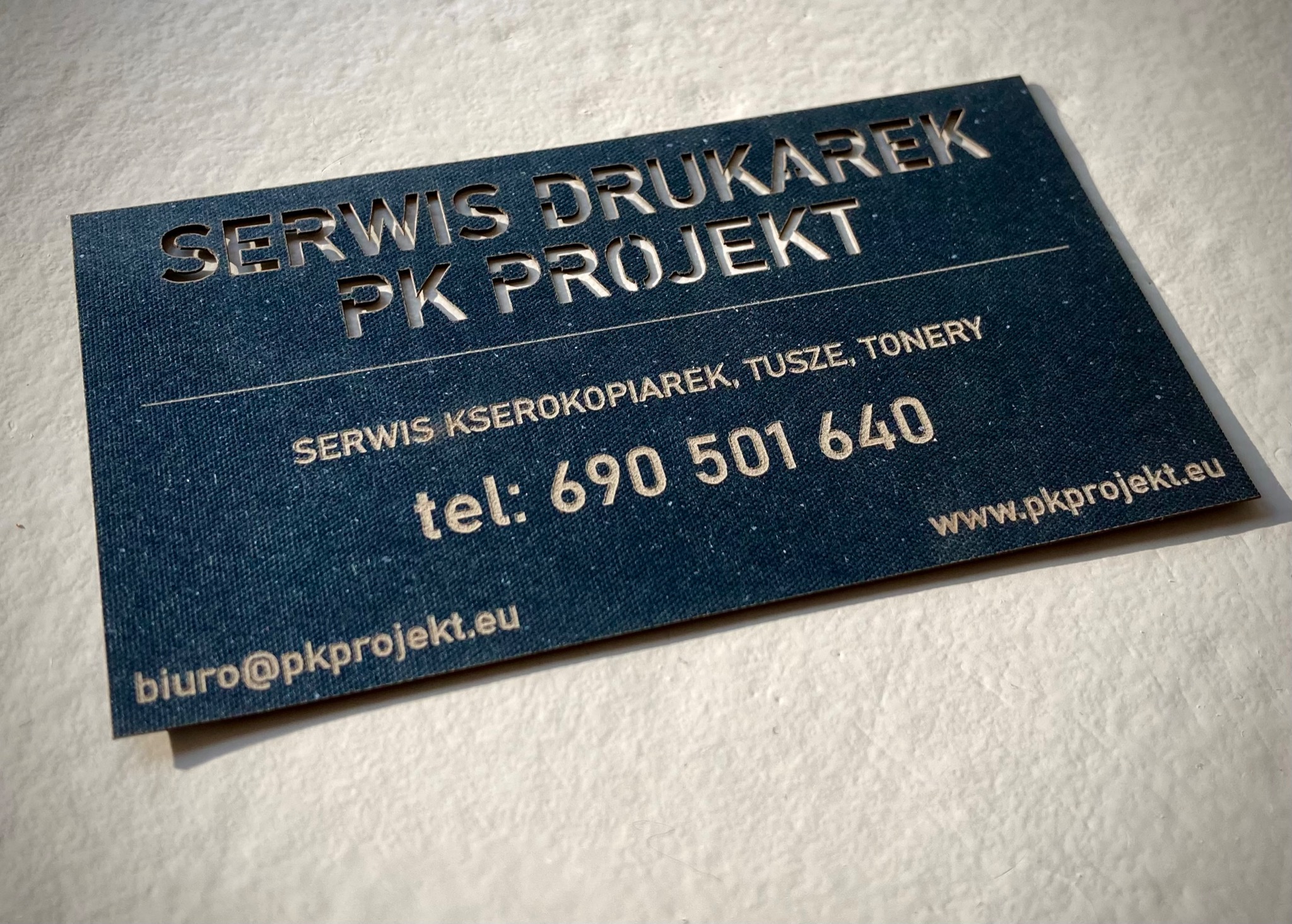 Wizytówka firmy 'Serwis Drukarek PK Projekt' z wyciętymi literami, oferującej serwis kserokopiarek, tusze i tonery, z numerem telefonu i adresem e-mail.