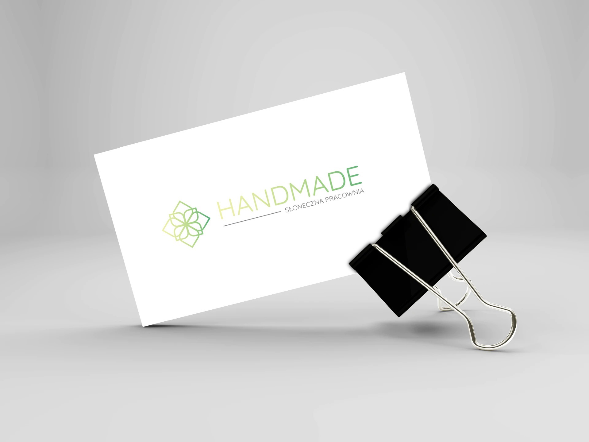 Wizytówka z zielonym logo kwiatowym i napisem 'HANDMADE Słoneczna Pracownia', przytrzymywana czarnym klipsem na jasnym tle.