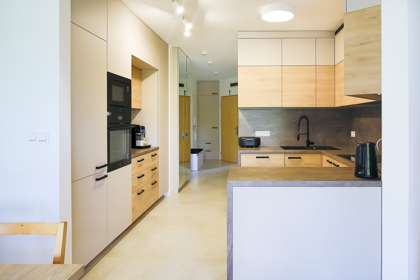 Nowoczesna kuchnia z beżowymi i drewnianymi frontami, czarnymi uchwytami, zabudowanym piekarnikiem i mikrofalówką, oraz wyspą kuchenną z szarym blatem, widok na przedpokój z lustrem.