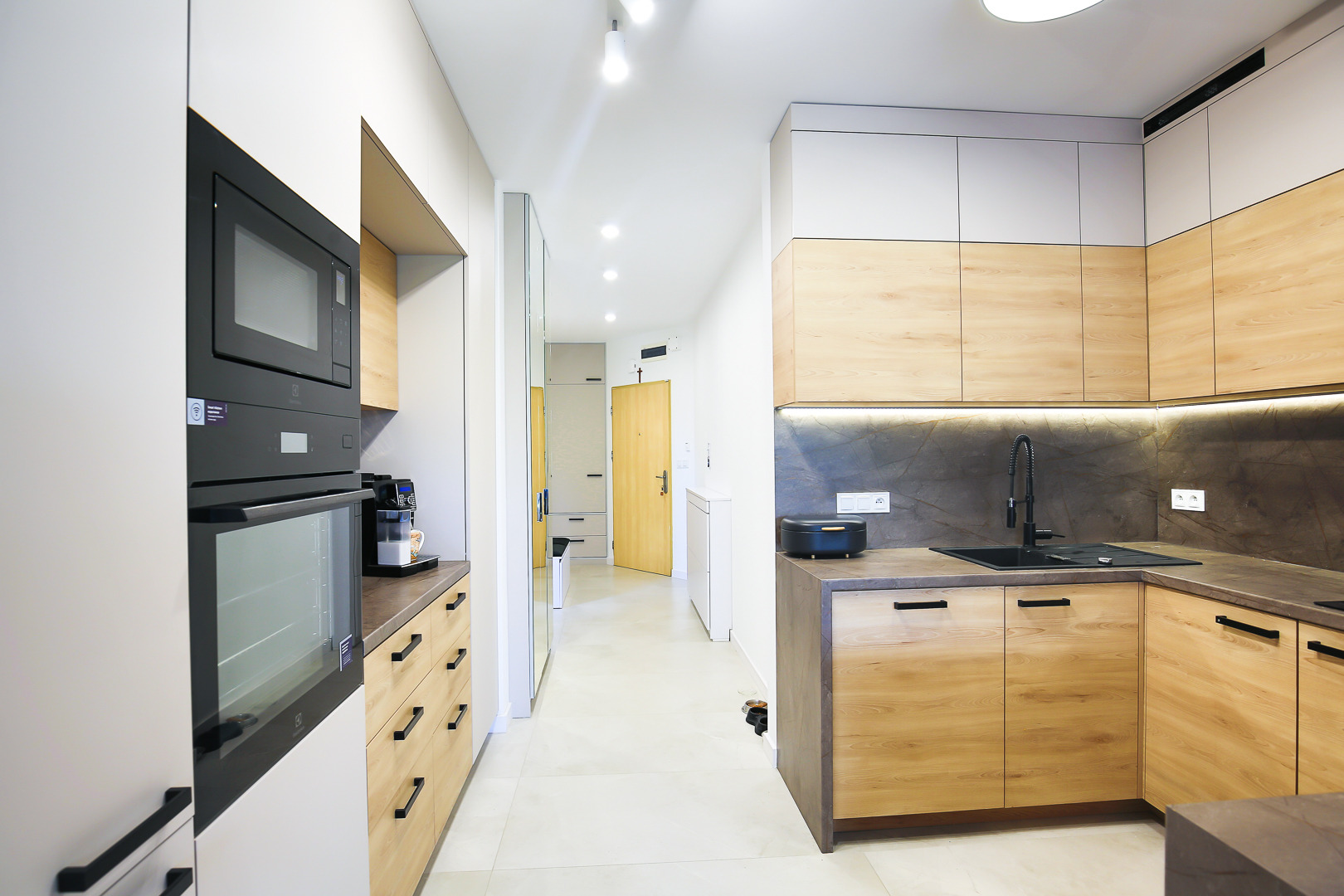 Nowoczesna kuchnia z drewnianymi szafkami i czarnymi elementami, w tle widoczny korytarz z lustrem i jasnymi drzwiami.