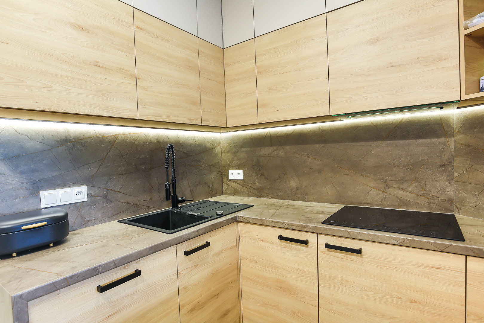 Nowoczesna kuchnia w jasnych odcieniach drewna z czarnymi akcentami: bateria, zlew i uchwyty, marmurowy panel nad blatem, oświetlenie LED pod szafkami.