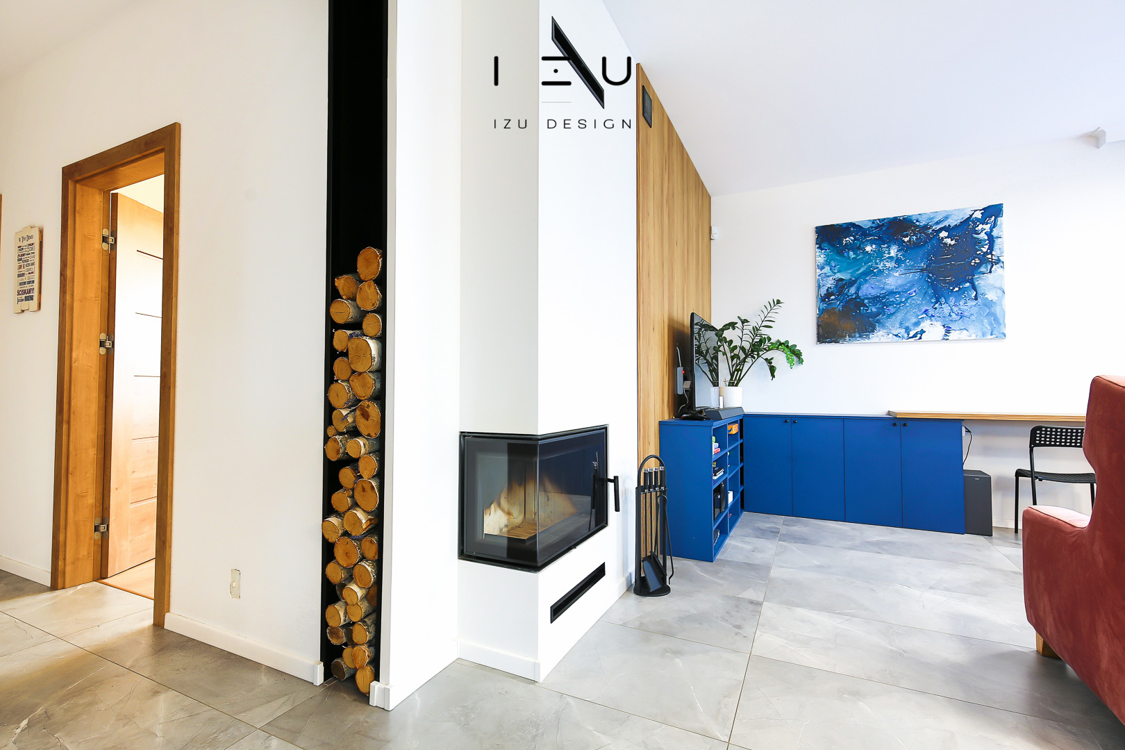 Nowoczesny salon z kominkiem narożnym, wnęką na drewno i niebieską zabudową meblową pod telewizorem, uzupełniony abstrakcyjnym obrazem w odcieniach błękitu.