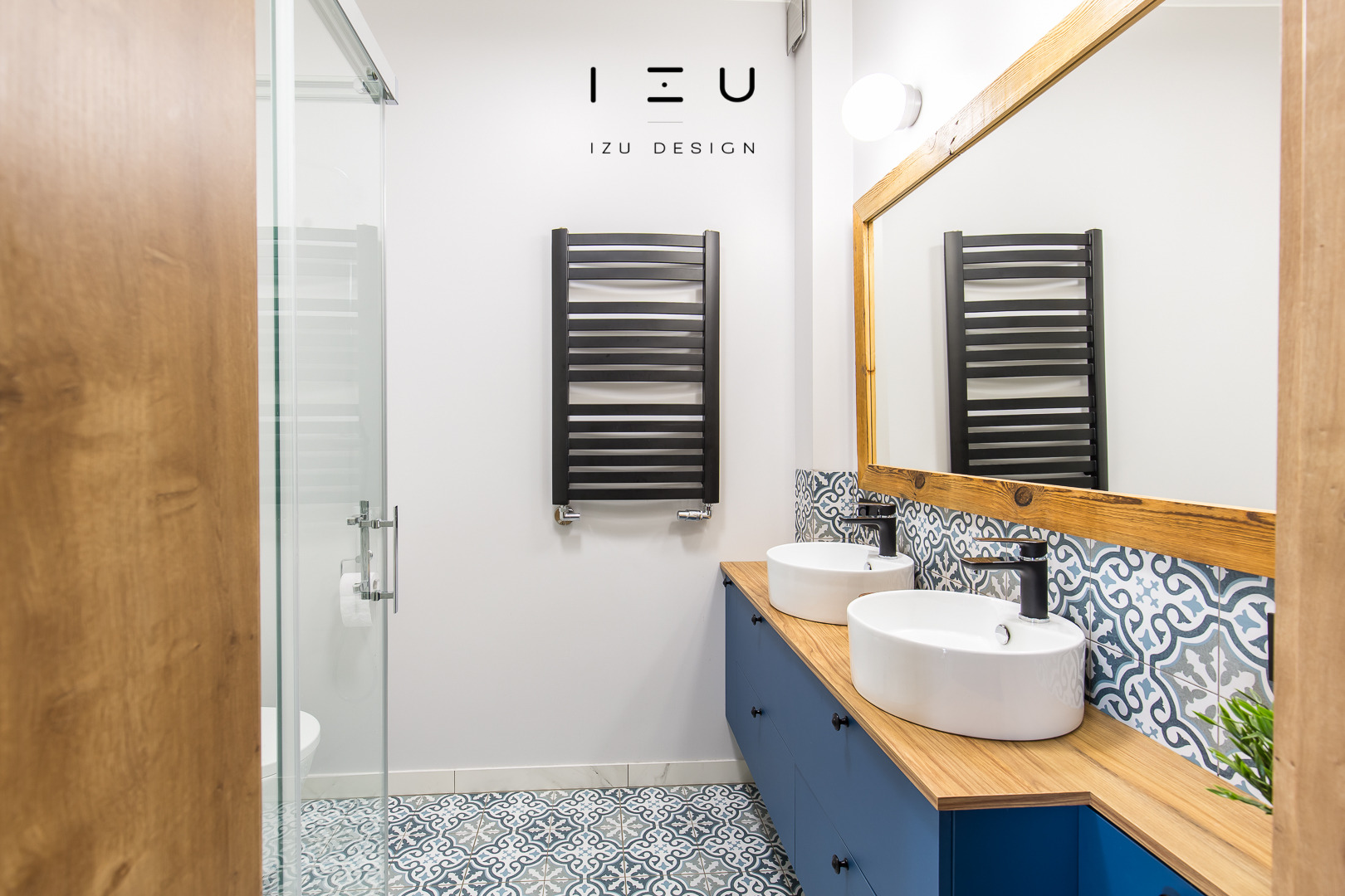 Nowoczesna łazienka z dwoma umywalkami nablatowymi, niebieską szafką, drewnianym blatem i lustrem w drewnianej ramie, czarny grzejnik drabinkowy na ścianie, kabina prysznicowa z przezroczystego szkła.