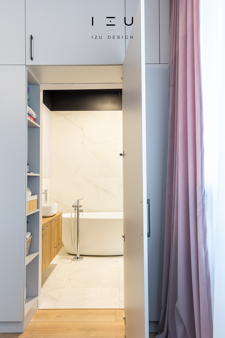 Nowoczesna łazienka widziana zza uchylonych drzwi szafy wnękowej, z białą wanną wolnostojącą, umywalką nablatową na drewnianej szafce, marmurowymi płytkami i pudrowo-różową zasłoną.