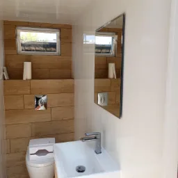 Nowoczesna, jasna łazienka w Koninie z drewnopodobnymi płytkami na ścianie, umywalką z szafką i lustrem. Widoczne okno i toaleta z pudełkiem nawilżanych chusteczek.