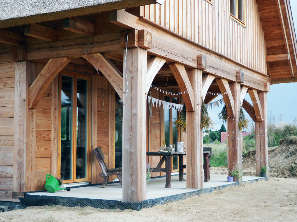 Dom jednorodzinny w Przechlewie z drewnianą elewacją, strzechą i werandą z dekoracjami. Widoczny zielony konewka przy drzwiach werandy.