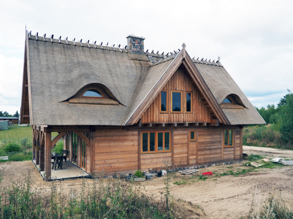 Drewniany dom jednorodzinny z dachem krytym strzechą i oknami w kształcie oka, zlokalizowany w Przechlewie, w trakcie wykańczania terenu.