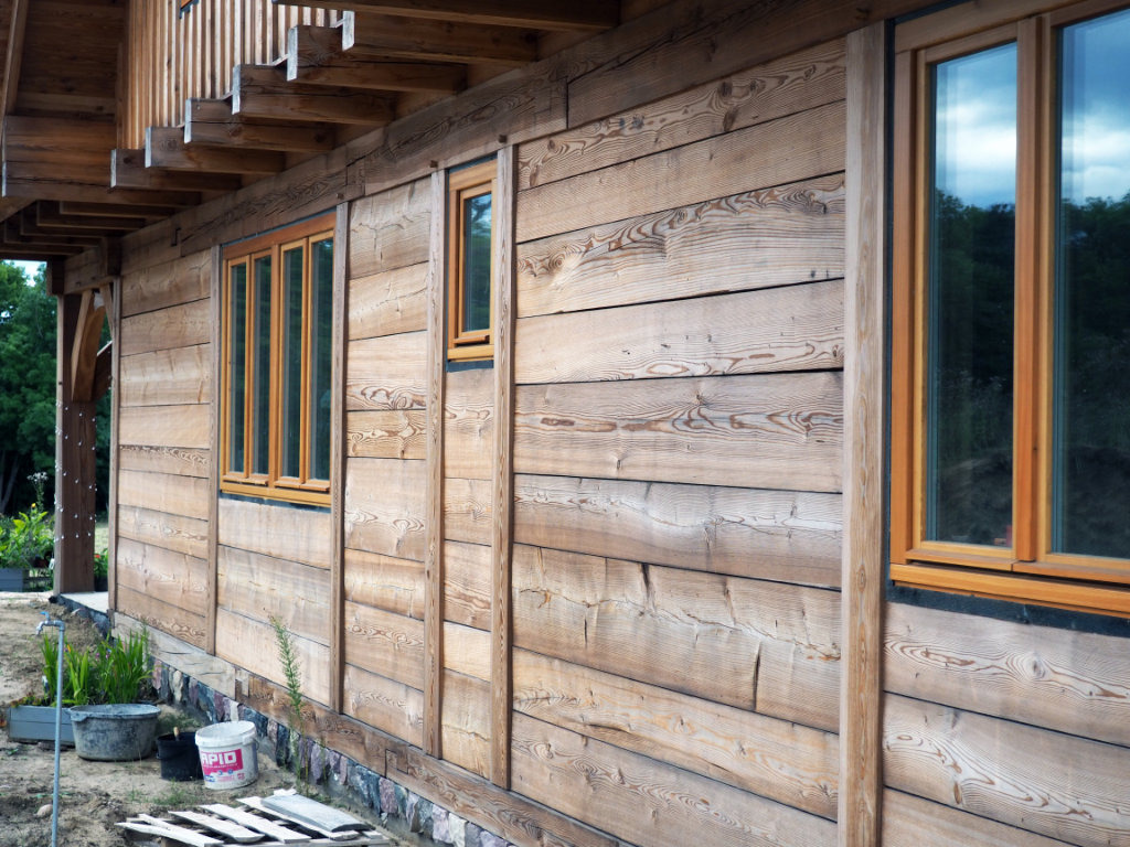 Drewniana elewacja domu jednorodzinnego z widocznymi oknami w drewnianych ramach i fragmentem balkonu, z detalami wykończenia i elementami ogrodu w tle.