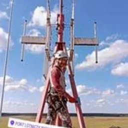 Pracownik w uprzęży na szczycie metalowej konstrukcji wieży telekomunikacyjnej, w tle błękitne niebo i odległy krajobraz.