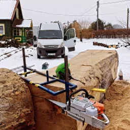 Przenośny tartak z piłą łańcuchową tnie duży, jasny pień drzewa na tle zimowego krajobrazu z dostawczym samochodem Iveco i domem w Mrągowie.