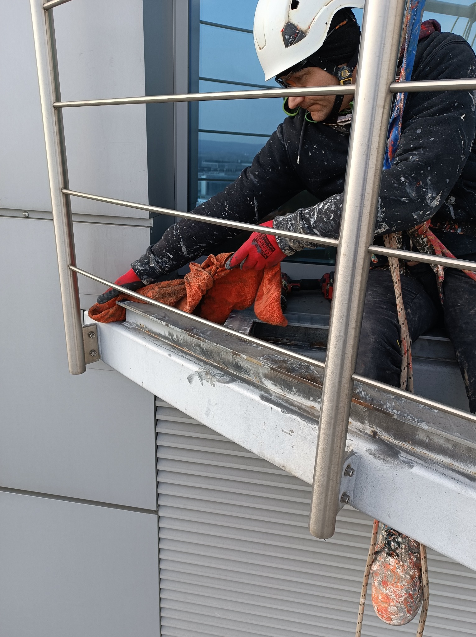 Pracownik w uprzęży, z kaskiem i w rękawicach roboczych, czyści szmatką metalową konstrukcję na wysokości, widoczny fragment elewacji budynku.