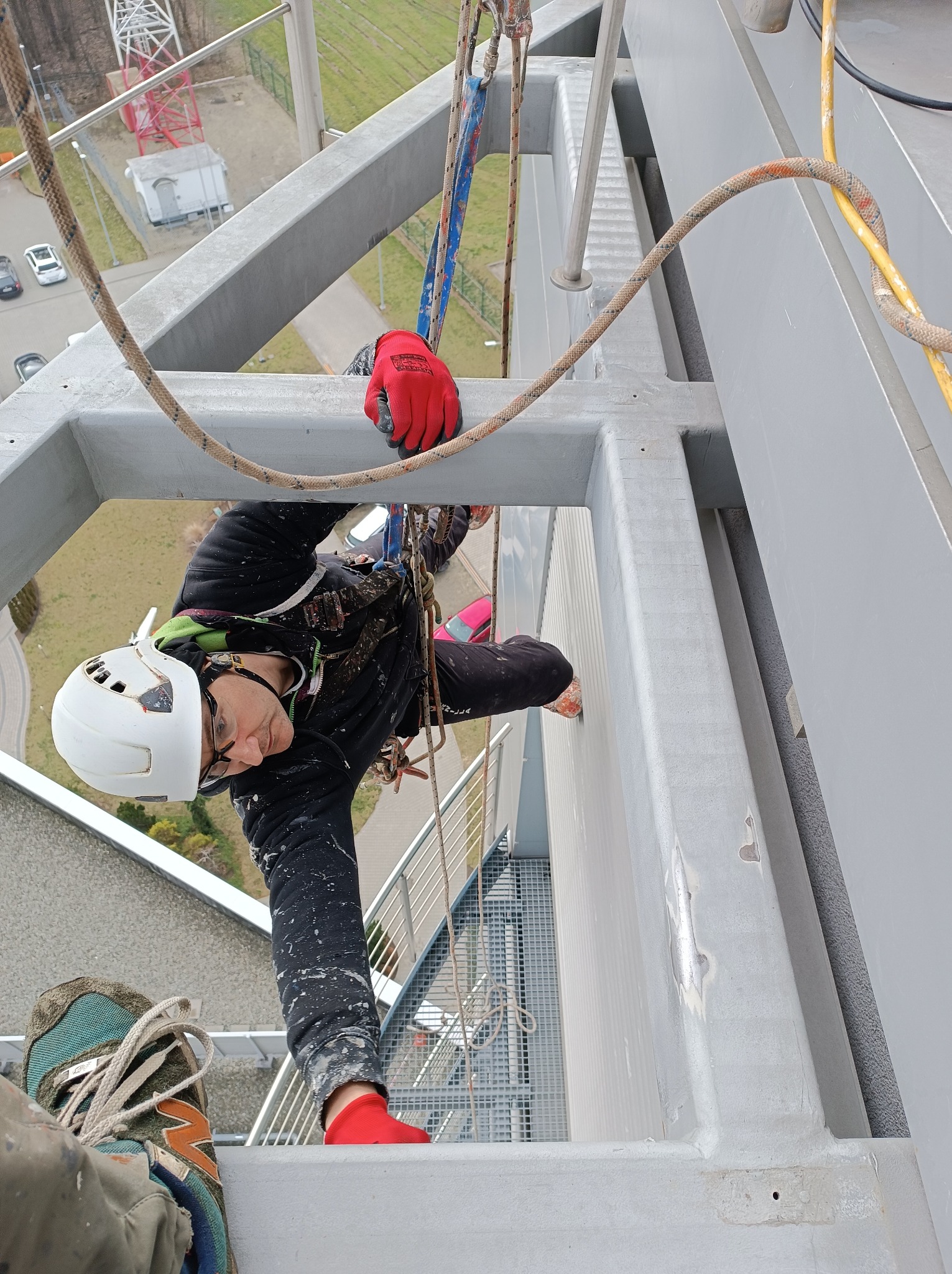Pracownik w kasku i okularach, zawieszony na linach, wykonuje prace na wysokości na metalowej konstrukcji, widoczne czerwone rękawice i skarpetki, w tle budynki i trawnik.