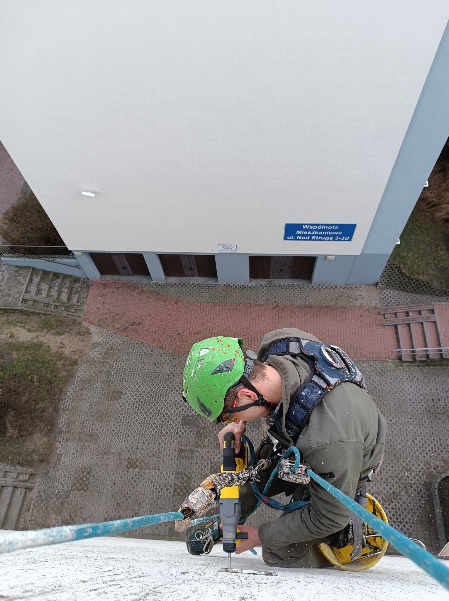 Pracownik w kasku i uprzęży, z perspektywy z góry, wierci w elewacji budynku w pobliżu tabliczki z adresem, zabezpieczony linami.