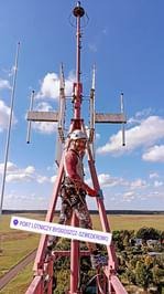 Pracownik w uprzęży na szczycie metalowej konstrukcji wieży telekomunikacyjnej, w tle błękitne niebo i odległy krajobraz.
