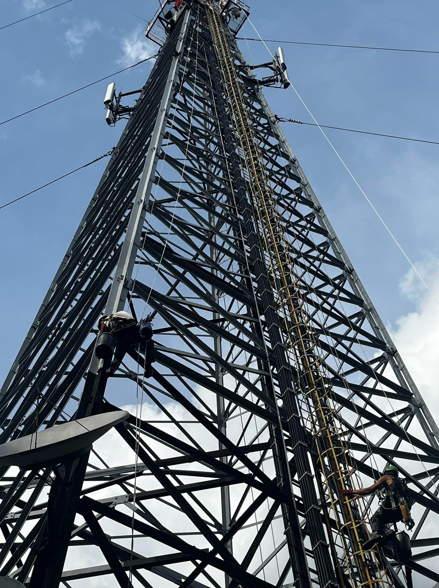 Dwóch pracowników na stalowej wieży telekomunikacyjnej podczas prac konserwacyjnych, widok z dołu na tle błękitnego nieba.