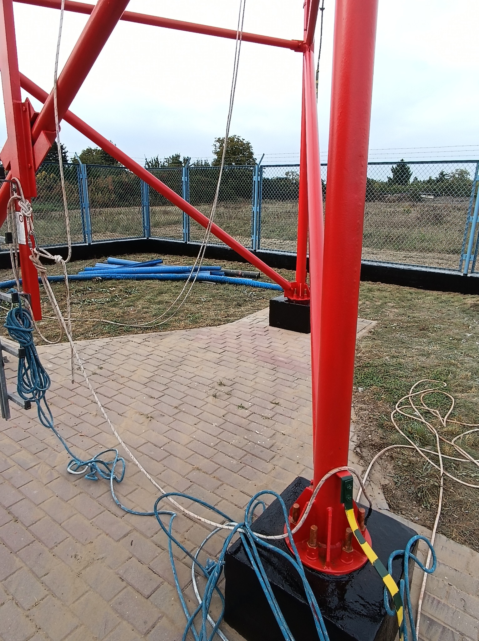 Czerwona konstrukcja stalowa z linami i rurami na zewnątrz, zabezpieczona ogrodzeniem z siatki.