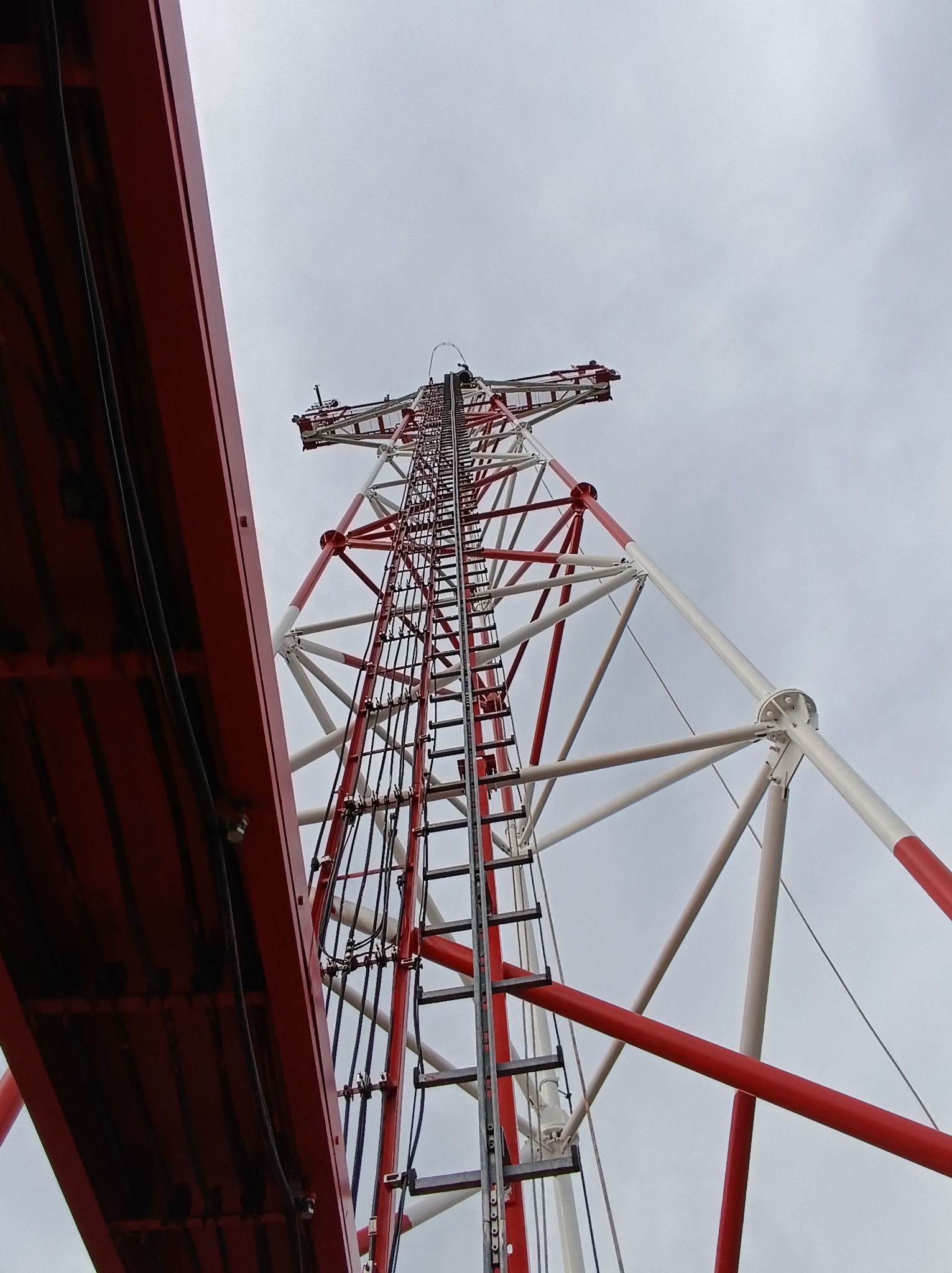 Wysoka, stalowa konstrukcja wieży telekomunikacyjnej pomalowana na biało i czerwono, widziana z dołu pod kątem, z drabinką prowadzącą na szczyt i fragmentem czerwonej platformy z kablami po lewej...