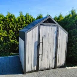 Drewniana szopa na narzędzia z metalową ramą i dachem, pomalowana na biało, z długim metalowym uchwytem, stojąca na kostce brukowej na tle zielonych tuji i błękitnego nieba.