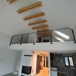 Wnętrze domu z antresolą zabezpieczoną czarną metalową balustradą. Widoczne drewniane belki stropowe, białe ściany, kominek i fragment kuchni w tle.