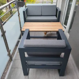 Nowoczesny balkon w Andrychowie z meblami ogrodowymi: grafitowy zestaw wypoczynkowy z drewnianym stolikiem i szklaną balustradą ze stali nierdzewnej.