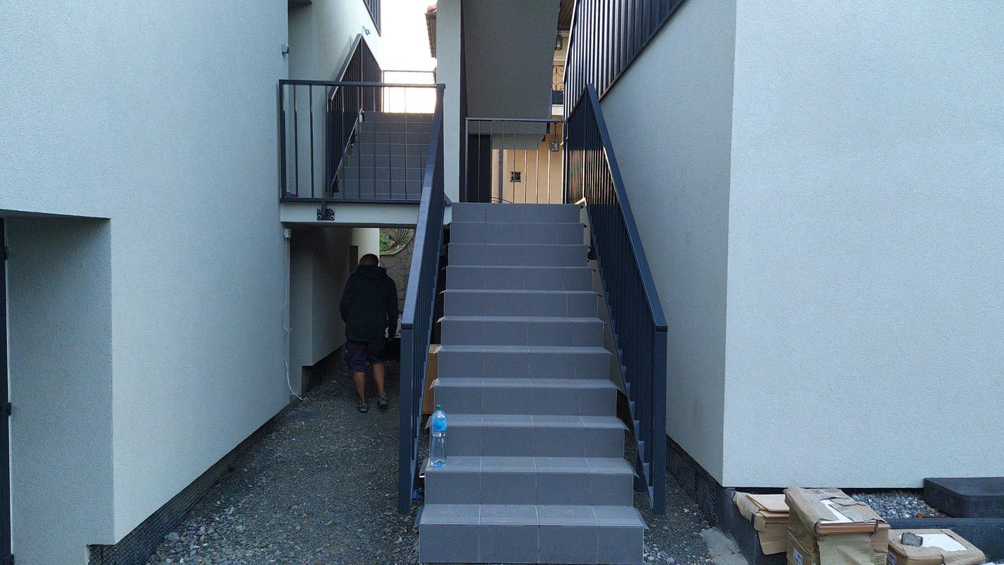 Zewnętrzne schody z szarych płytek z czarną balustradą, prowadzące na wyższy poziom budynku, obok zaparkowana butelka wody, w tle osoba wchodząca pod schody.