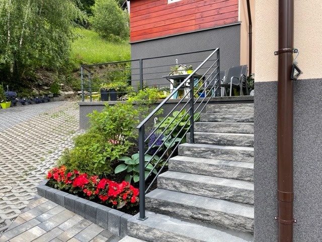 Zewnętrzne schody z szarego kamienia prowadzące do domu, zabezpieczone prostą, czarną balustradą, otoczone rabatą z czerwonymi kwiatami i zielenią, z widokiem na trawnik i drzewa w tle.