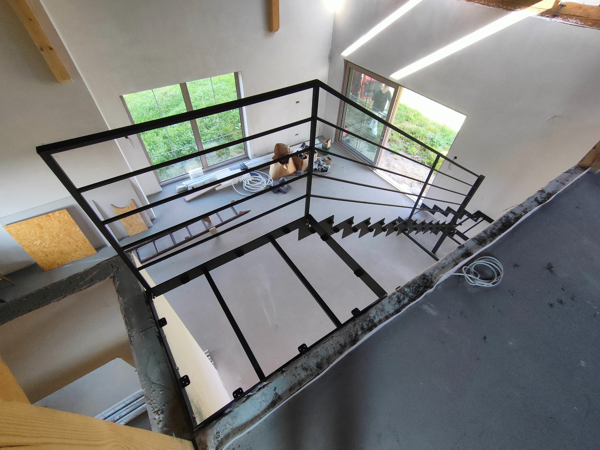 Wysoki kąt widzenia na surowe schody z czarną metalową balustradą w nowym domu, widoczne narzędzia i materiały budowlane, okna z widokiem na zieleń.