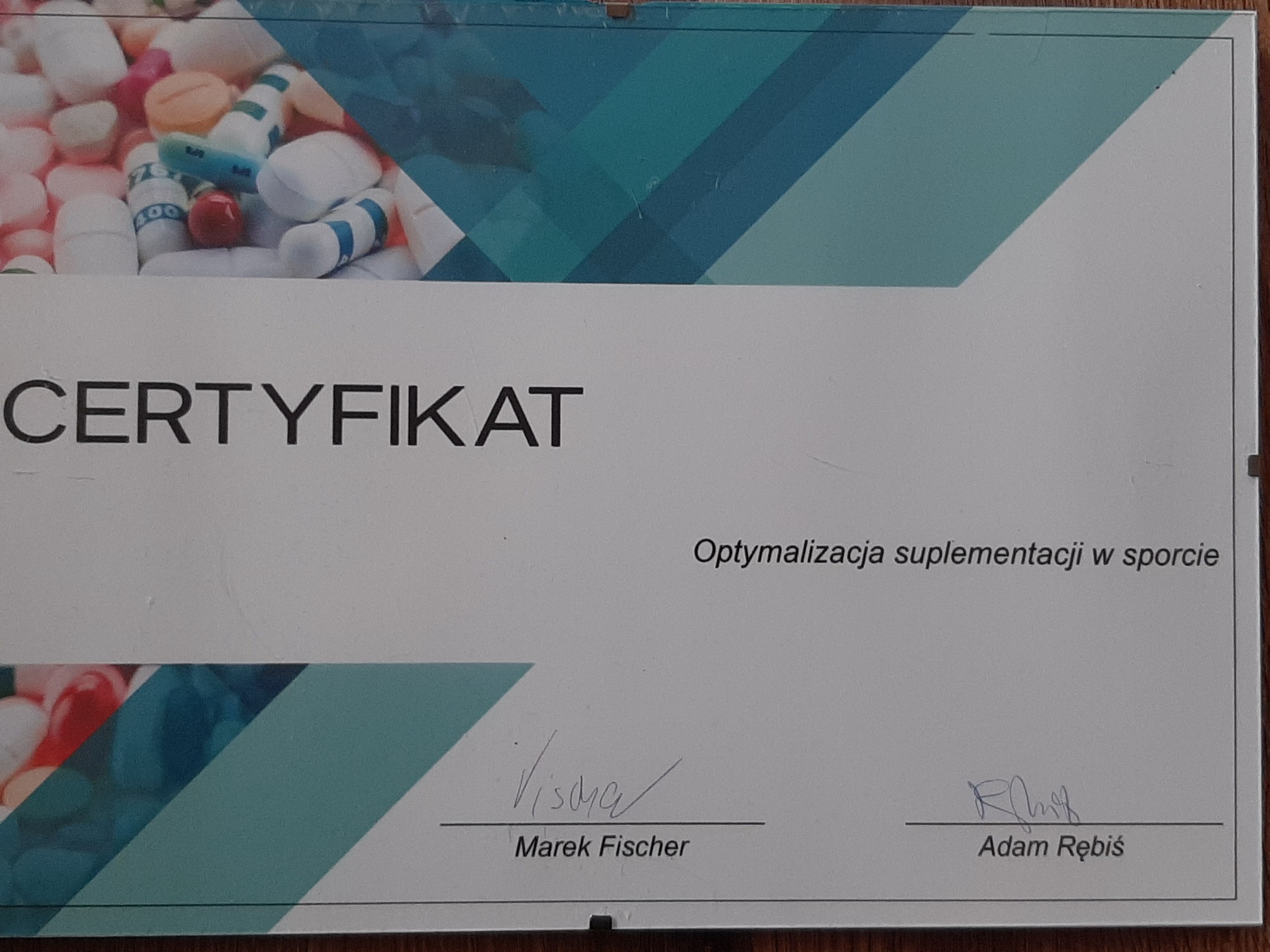 Certyfikat optymalizacji suplementacji w sporcie, z widocznymi podpisami Marka Fischera i Adama Rębiś, tło z różnokolorowymi pigułkami i kapsułkami.
